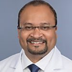 Head shot of man with trim beard, glasses, wearing white shirt, dark tie and physician's white coat