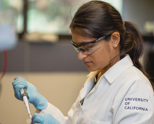 Researcher in a UC‑branded lab coat and safety gear pipetting liquid into a small container at a laboratory.