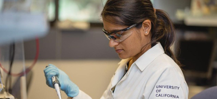 Researcher in a UC‑branded lab coat and safety gear pipetting liquid into a small container at a laboratory.
