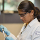 Researcher in a UC‑branded lab coat and safety gear pipetting liquid into a small container at a laboratory.