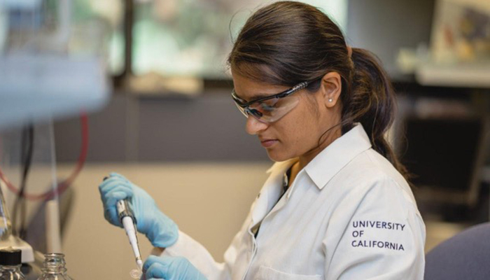 Researcher in a UC‑branded lab coat and safety gear pipetting liquid into a small container at a laboratory.