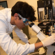 A researcher in a white coat and safety glasses examines a sample under a microscope in a laboratory.