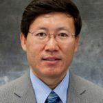 Head shot of man with black hair, glasses, grey suit and blue tie